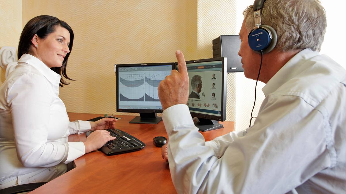 Starší muž se sluchátky při vyšetření sluchu, vedle něj audioložka u počítače