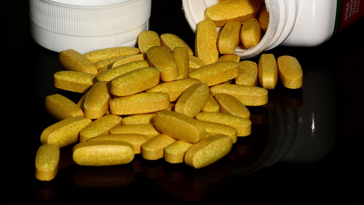 Žluté tablety vitaminů skupiny B vysypané z bílé lahvičky na černé lesklé ploše