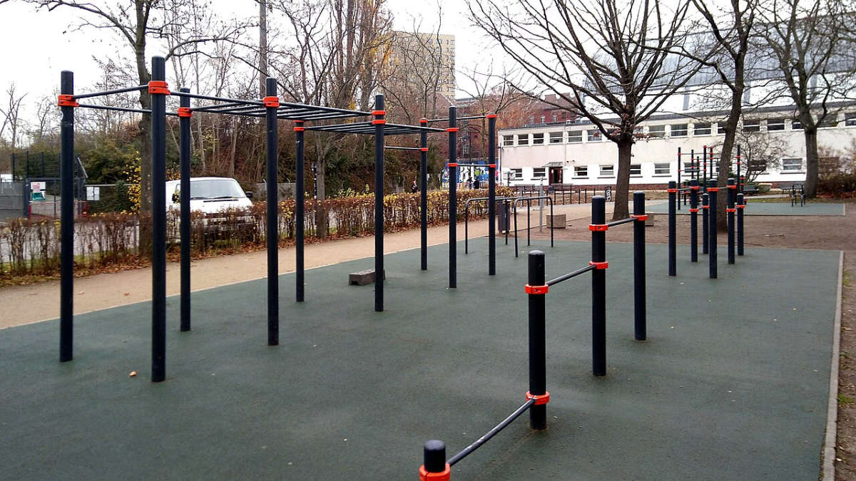 Venkovní hřiště na street workout s hrazdami a bradly na pryžovém povrchu, v pozadí stromy a budova