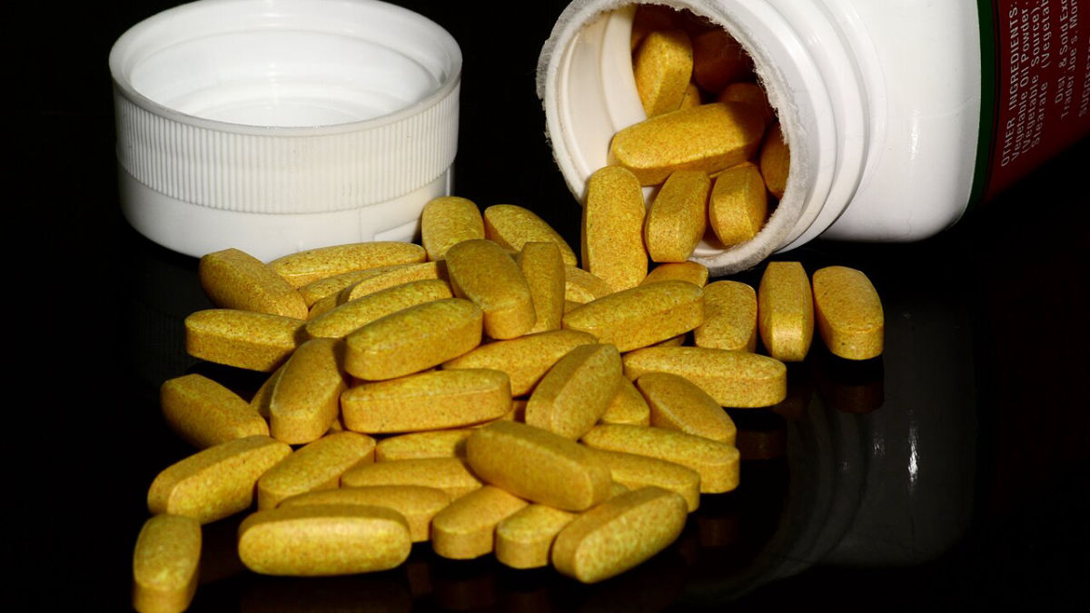 Žluté tablety vitaminů skupiny B vysypané z otevřené lahvičky na tmavém podkladu