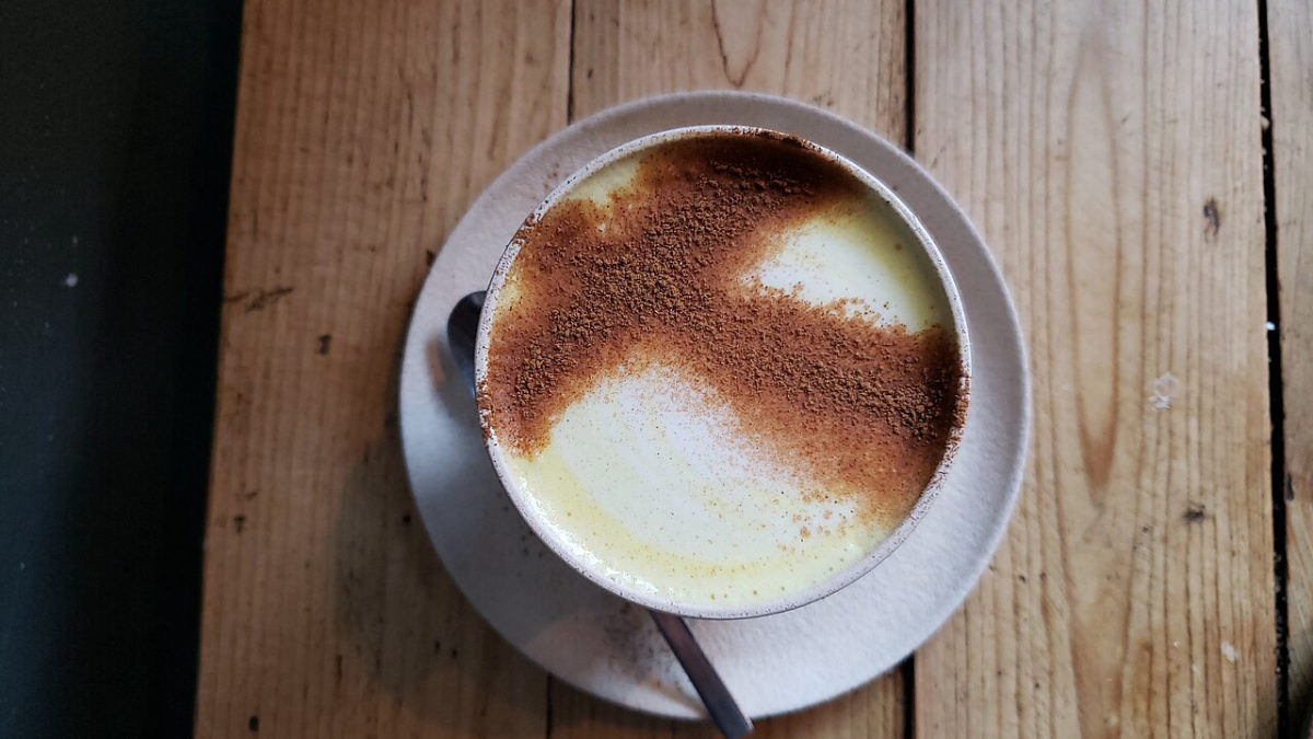 Hrnek kurkumového latte s mléčnou pěnou posypanou kořením na dřevěném stole, pohled shora